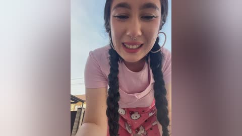 Video of a smiling, fair-skinned woman with long braided hair, wearing a pink T-shirt and a red skirt with Hello Kitty pattern, indoors.