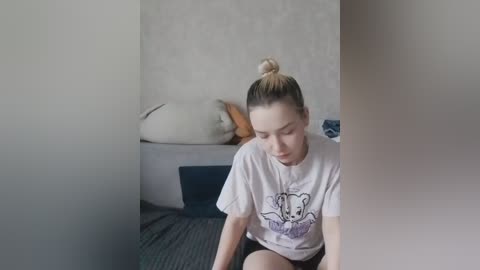 Video of a young woman with blonde hair in a bun, wearing a white T-shirt with a cartoon character, sitting on a bed with gray bedding, surrounded by a blurred, white wall.
