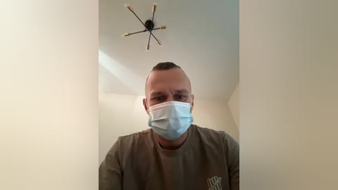 Video of a man with a bald head, wearing a light blue face mask and a gray t-shirt, standing in a beige room with a modern chandelier.