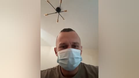 A video of a bald man with a light complexion wearing a light blue medical mask indoors, standing against a white wall with a modern, multi-arm chandelier hanging overhead.