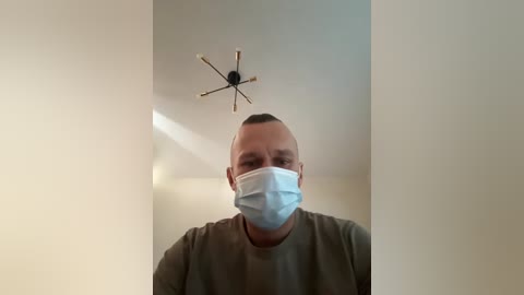 Video of a bald, light-skinned man with a surgical mask and a green T-shirt, standing indoors against a white wall, with a decorative starburst light fixture above.