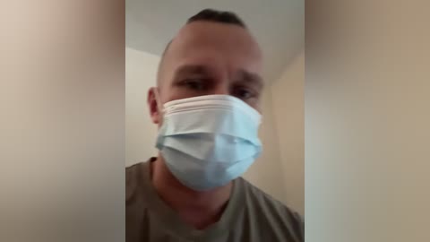 A blurry video of a man in a light brown T-shirt, wearing a blue surgical mask, with short dark hair. The background features beige walls and a ceiling.