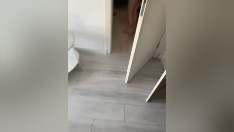 A video of a narrow, dimly lit hallway with light gray tiled flooring. A white doorframe and wall are visible, with a partially open door leading to a darker, indistinct room. The scene has a minimalist, slightly eerie atmosphere.