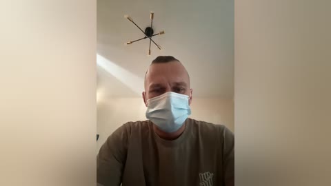 A video of a bald, middle-aged man with a light complexion wearing a blue surgical mask, a gray T-shirt, and standing in a dimly lit, beige-walled room with a modern, starburst ceiling light.