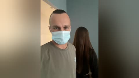 Video of a man in a beige T-shirt, wearing a light blue surgical mask, standing in a dimly lit room with a woman with long, straight brown hair.