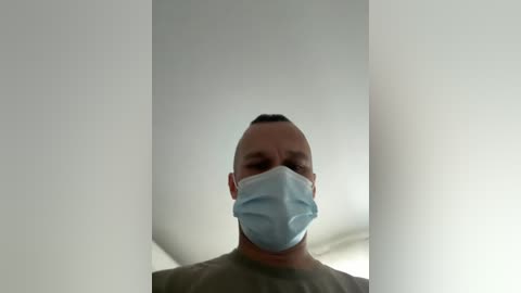 A video of a bald, light-skinned man wearing a blue medical mask, standing in a white-walled room with a ceiling light.