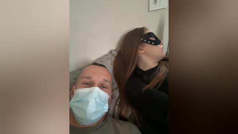 Video of a man in a hospital bed, wearing a mask, next to a woman in a black mask and black top, both looking to the right.