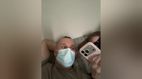 A video of a bald, light-skinned man in a gray shirt, wearing a light blue mask, taking a selfie with a pink iPhone. The background shows a beige wall and a white pillow.