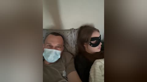 Video of a man and woman in a dimly lit room, both wearing surgical masks. The woman has long brown hair and a black mask with a cutout for the eyes.