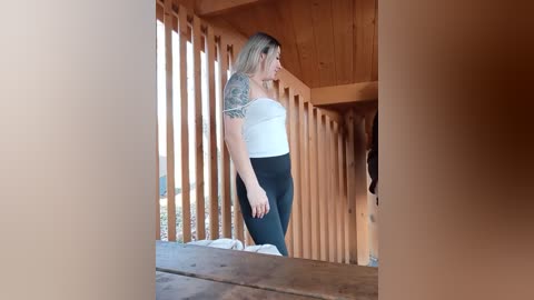 Video of a blonde woman with a tattooed arm, wearing a white tank top and black leggings, standing on a wooden deck with vertical beams.