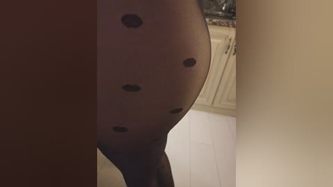 Video of a close-up of a person's thigh, clad in sheer black stockings adorned with small, black polka dots. Background features a beige wall and a partially visible kitchen cabinet.