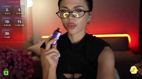 A video of a young woman with fair skin, dark hair, and glasses, wearing a black dress, holding a phone, against a blurred, dimly lit background.
