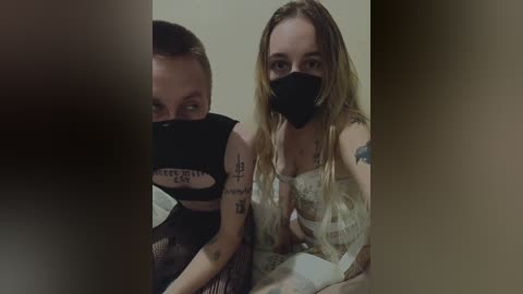 Video of two women, one with blonde hair and a black face mask, the other with a short haircut, both wearing black crop tops, sitting close together indoors.