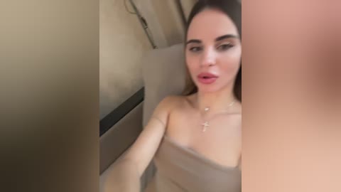 A video of a young woman with light skin and straight brown hair, wearing a beige towel, posing in a bathroom with beige walls and a white sink.