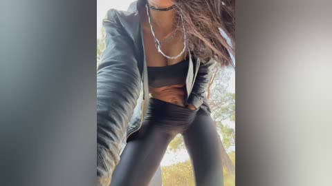 Video of a woman with medium skin tone, wearing a black bra, black leggings, and a black leather jacket, posing outdoors with a blurred background.