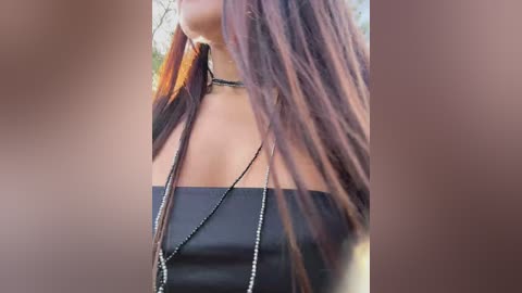 Video of a woman with long, straight brown hair, wearing a black strapless top and a long black beaded necklace, in a blurred outdoor setting.