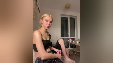 Video of a slim, blonde woman with fair skin, sitting barefoot on a sofa, wearing a black sports bra and patterned leggings. Background features a minimalist room with a woven ceiling lamp, window, and scattered furniture.