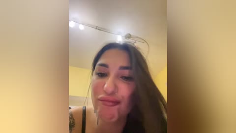 A video of a young woman with long brown hair, fair skin, and a nose ring, smiling, taken indoors under fluorescent lighting, with a yellow wall background.