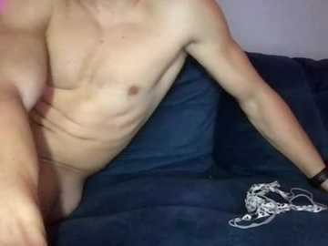 Video of a nude, light-skinned, fit man with a lean physique, sitting on a dark blue couch, his left arm resting on the armrest, with a silver chain and a black wristband on his wrist.