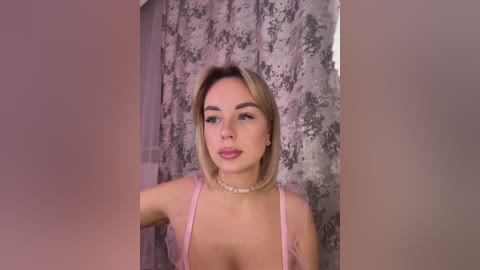 A video of a fair-skinned woman with straight blonde hair, wearing a pink bralette and a pearl necklace, standing against a floral-patterned grey curtain. She has a subtle smile.