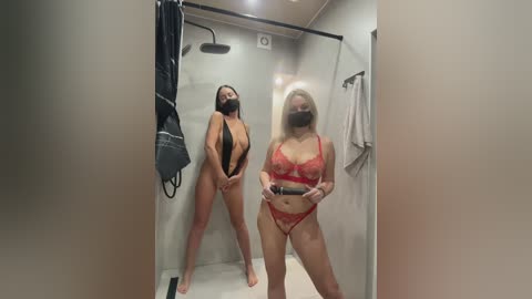 Video of two women in a modern, beige-tiled shower. One, with long black hair, wears a black bodysuit, and the other, blonde, in red lace lingerie, holds a camera.