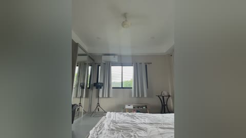 Video of a modern, minimalist bedroom with a white bed, beige walls, and a large window with white curtains. A ceiling fan and a small table with a lamp are visible.