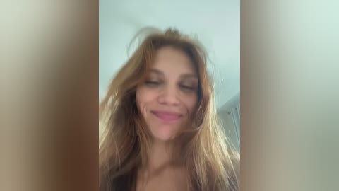 Video of a smiling, fair-skinned woman with long, wavy blonde hair, wearing a dark top, set against a blurred, light-colored background.