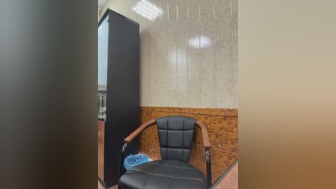 Video of a small, sparsely furnished room with a black leather chair, wooden paneling, and a closed door.