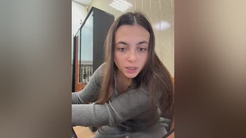 Video of a young woman with long, straight brown hair, wearing a gray ribbed sweater, sitting at a wooden table in a dimly lit, beige-paneled room, with blurry out-of-focus people in the foreground.