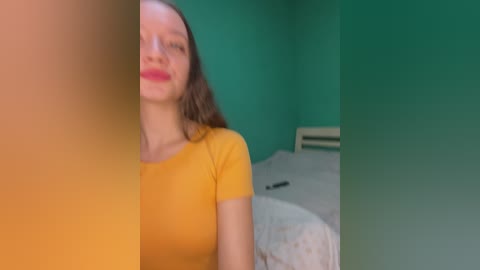 Video of a young woman with long brown hair, wearing a yellow shirt, standing in a teal-painted room with a bed and white bedding in the background.