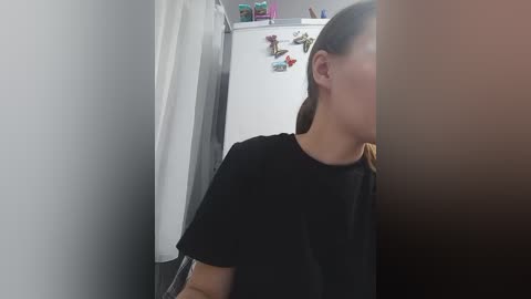 Video of a woman with light skin and straight, dark hair, partially visible, wearing a black t-shirt, standing in a narrow hallway with a white door and a gray curtain.