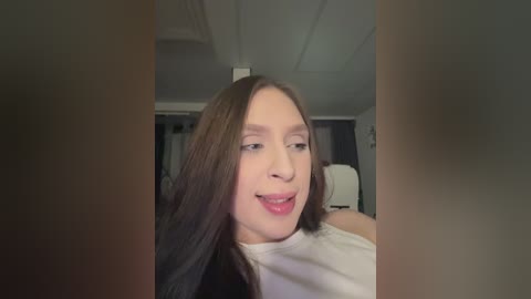 Video of a smiling young woman with long brown hair, wearing a white top, in a dimly lit room with a white ceiling and dark curtains.