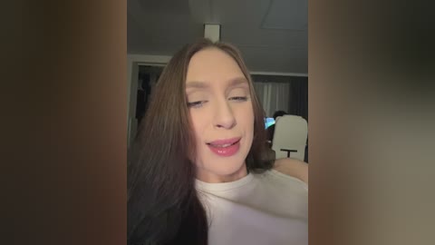 Video of a Caucasian woman with long, straight brown hair, wearing a white top, smiling and sticking out her tongue in a playful manner. Background shows a dimly lit room with a white chair and blurred details.