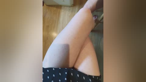 A video shows a woman's legs in black shorts with white dots, lying on a wooden floor, with a beige chair and part of a beige couch in the background.