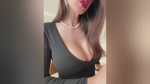 Video of a woman with long brown hair, wearing a black one-shoulder dress, revealing ample cleavage, and bright red lipstick. She has a pearl necklace and a subtle blush.
