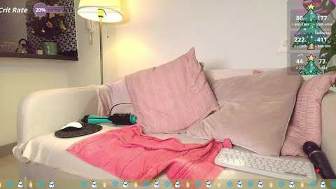 Video of a cozy, cluttered living room with a beige couch, pink cushions, a white keyboard, a green mouse, a pink blanket, and a Christmas tree in the background.