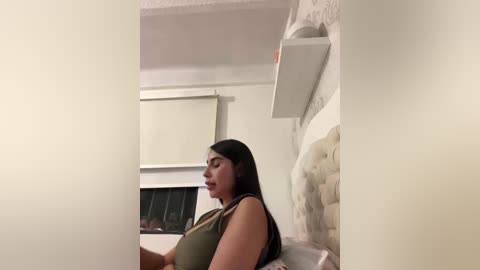 A video of a Latina woman with long black hair, medium build, and medium skin tone, wearing a green tank top, lying on a beige couch in a dimly lit, modern living room with white walls and a white ceiling.