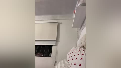 Video of a dimly lit, cluttered bedroom with a white bed adorned with a red heart-patterned duvet, a white wall-mounted air conditioner, and a white shelf above it.