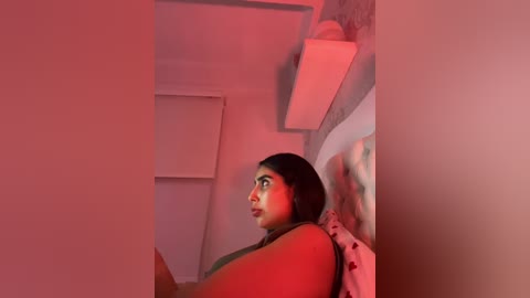 Video of a young woman with medium brown skin and long black hair, wearing a sleeveless top, sitting on a bed in a dimly lit room with red lighting.