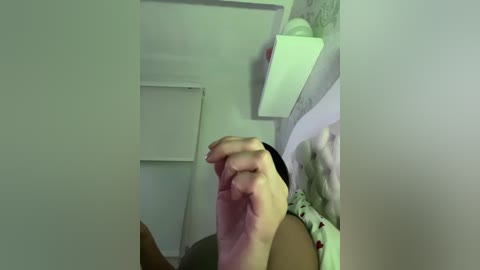 Video of a close-up hand holding a smartphone, seen from a low angle, with a bed in the background featuring floral-patterned bedding and a white shelf. The image has a soft, slightly blurred focus.