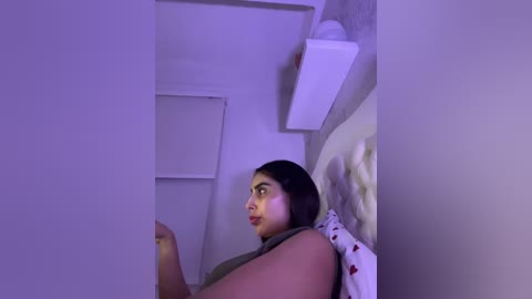 Video of a young woman with dark hair, medium complexion, and a curvaceous physique, sitting on a bed in a dimly lit bedroom with a white wall, white pillow, and a small lamp.
