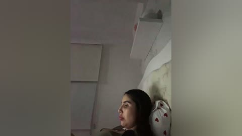 A video shows a young woman with long dark hair, lying on a bed with white bedding and heart-patterned pillowcases, in a dimly lit room with white walls and a white shelf.