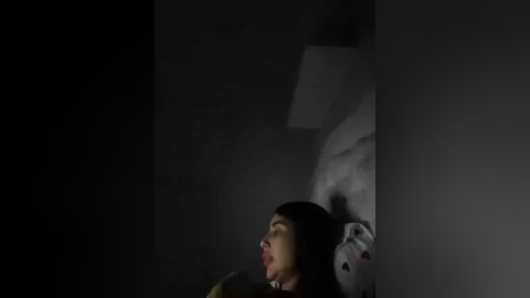 A dimly-lit video of a woman with long, dark hair and pale skin, lying on a bed with white sheets. She has a distressed expression, with her mouth open, and her eyes closed. A stuffed animal, possibly a bear, is partially visible on the pillow beside her.