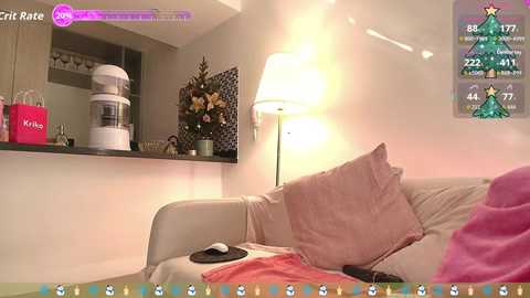 A cozy living room scene with a beige sofa, a pink throw, a white lamp, and a Christmas tree on the wall.