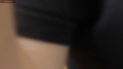 A blurred video of a dark-skinned woman wearing a black outfit, standing in a dimly lit room with a beige wall in the background.