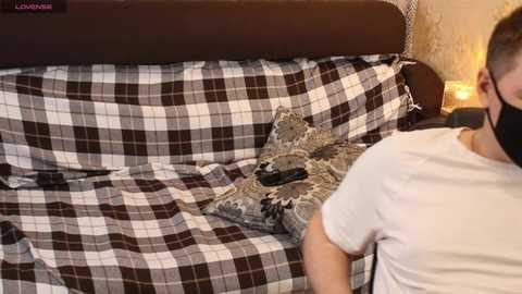 A video of a man wearing a white t-shirt and a face mask, standing beside a plaid-covered sofa with a decorative pillow.