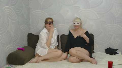 Video of two women, one blonde, one platinum blonde, sitting on a bed, wearing white and black masks, white shirts, and short shorts, with a gray-patterned wallpaper background.