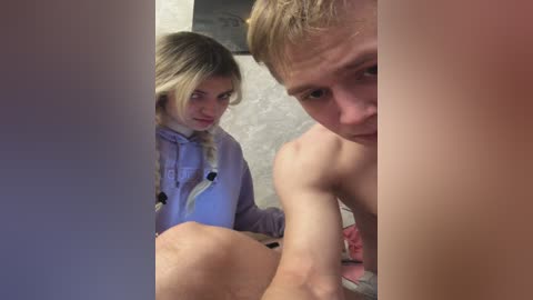 Video of a blonde woman in a lavender hoodie peering through a doorway at a shirtless man with short hair, looking concerned.