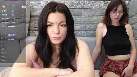 Video of a woman in a pink tank top, sitting on a bed, and a woman in a black crop top and red plaid skirt, both looking at a smartphone.