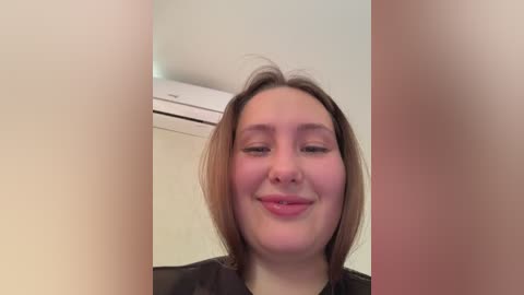 Video of a smiling, light-skinned woman with straight, shoulder-length brown hair, wearing a black top. Background shows a beige wall and a white air conditioning unit.